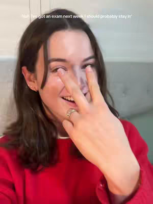 A young woman with long, dark hair holds up three fingers in front of her face looking at the camera like she is counting. She is wearing a red sweater, with a ring on her left hand. The background mostly shows the wall.