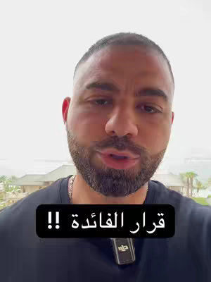 A man with a beard is looking at the camera, in front there is a black rectangle with white text, behind him there is a window showing a seascape