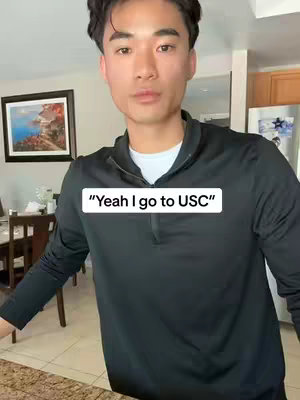A young Asian man stands in a kitchen setting, wearing a black quarter-zip shirt over a white undershirt. Behind him are simple kitchen cabinets and a framed picture. A white text overlay reads, "Yeah I go to USC".