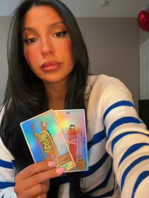 A woman is holding two tarot cards. She is wearing a white sweater with blue stripes. Her long, dark hair frames her face. She is looking at the camera with a neutral expression. The background is a plain wall with a red shape, possibly a decoration.