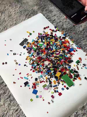 A top-down shot shows a white surface covered in a large assortment of colorful LEGO bricks scattered around. A smartphone is partially visible, as if taking a picture of the LEGOs. The background is a light, textured surface.