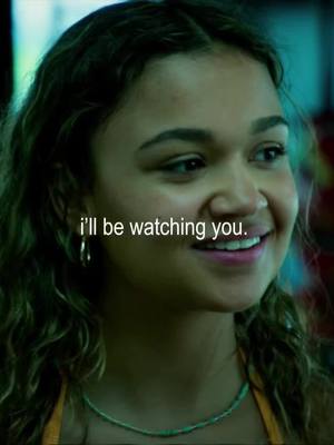 A close-up of a young woman with long, wavy hair, smiling with visible joy. She has a tan skin tone and is wearing hoop earrings and a necklace. The background is a blurred mix of colors, suggesting an interior setting with artistic lighting, and text overlayed on the image that says 'I'll be watching you'.