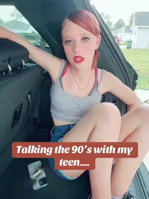 A young woman sits in the open back of a vehicle with a text overlay. She has red hair and is wearing a tank top and shorts. Her expression is one of slight amusement.