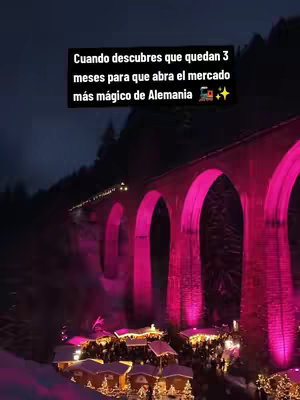 A nighttime shot of a market below a lit-up stone bridge. The bridge is vividly illuminated on its arches in pink, and the market stalls below are lit with warm, inviting lights, with a dark night sky above and some trees on the mountain behind the bridge.