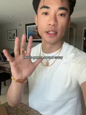 A man is in a room, with a hand in front of his face. He is wearing a white t-shirt and gold jewelry. Text appears to be overlaid on his hand.