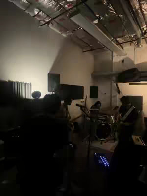 An indoor shot of a band performing on a stage. The lighting is dim, primarily from stage lights, with a focus on the band members and their instruments.