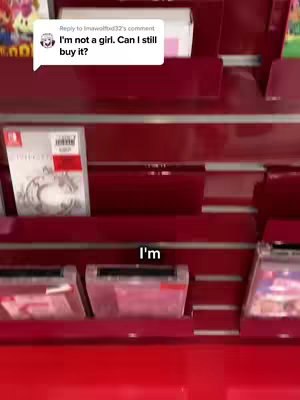 A person is filming inside a retail video game store, the image shows shelves holding some products. The overlay text in a chat bubble states: 'I'm not a girl. Can I still buy it?'