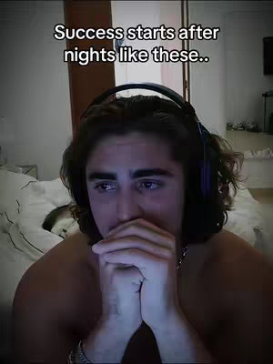 A young man sits on a bed, appearing contemplative with his hands clasped in front of his face. He's wearing a black headset, and the background suggests a bedroom setting with a window and possibly a computer screen. The overall color scheme is somewhat muted, with the lighting being dim and creating a sense of introspection.