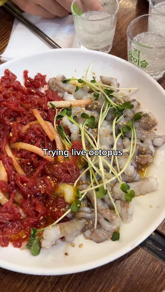 A close-up shot of a white plate filled with a variety of foods, including a portion of what appears to be raw, chopped red meat with thin orange strips and some greens such as sprouts. To the right is a serving of what appears to be live octopus, identifiable by small, translucent bodies and tentacles. The background is blurred, but suggests a dining setting with clear glasses containing a light-colored liquid.