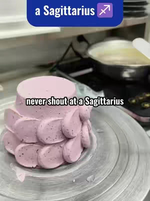 A close-up shot of a cake decorated with a pink frosting flower design on a rotating surface, likely in a kitchen setting, with text overlays stating 'a Sagittarius' and 'never shout at a Sagittarius'.