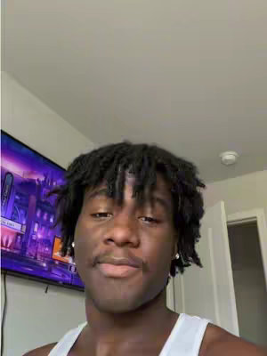 A young man with dark hair and a white tank top is centered in a selfie-style shot. A large TV screen displaying a blurry image is partially visible in the top left corner. The background features a light-colored wall and a door frame. The man is positioned close to the camera, with his face in focus and a neutral but engaged expression. He has a slight mustache and small earrings.