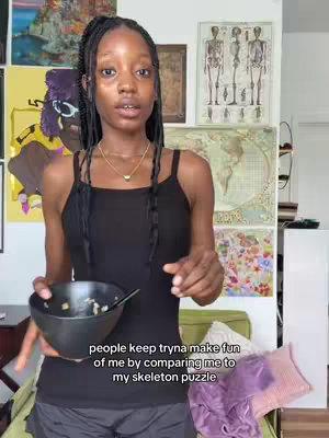 A young woman with long, dark braided hair stands indoors, holding a dark bowl with food and a small spoon in her left hand. She is facing the camera and wearing a black tank top. The background includes various artwork, posters, and a small green sofa.