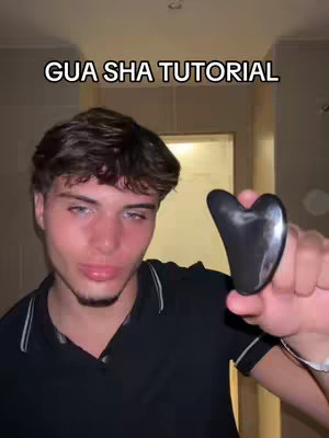 A young, fair-skinned man with short, styled brown hair is holding a black gua sha tool in his right hand. He is looking towards the camera. The background is a nondescript interior, likely a bathroom, with a door partially visible. The text overlay reads "GUA SHA TUTORIAL" in all caps.