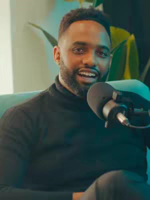 The cover image features a man with a dark beard and short, styled hair, appearing to be in a podcast setting. He wears a black turtleneck and smiles while looking slightly to the side of the camera. A microphone is positioned close to his mouth, indicating the recording of a podcast. The background has a blurred effect with hints of greenery and soft lighting, contributing to a professional studio look.
