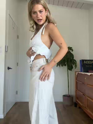 A woman stands indoors, facing the camera at a slight angle. She wears a white halter top and light-colored jeans. Her blonde hair is styled with a slight wave, and she is speaking. The background includes a door frame and a houseplant.