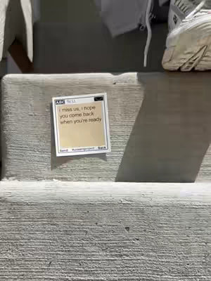 A square sticky note with text is adhered to what looks like a concrete surface. The text note has a light beige background with a rectangular white border, and a digital interface is on the top left with the logo "ABC Tv 11". The text on the note reads, "I miss us, I hope you come back when you're ready". Parts of a shoe, along with shadows and sunlight, are present in the frame.