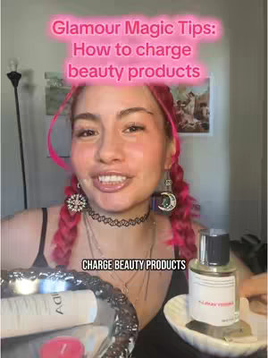 A face-centered shot featuring a woman with pink hair and a smiling expression. The background is slightly blurred, with hints of a room setting and objects like beauty products in the foreground.