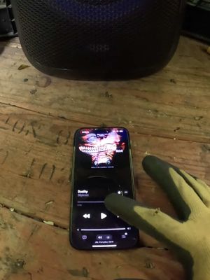 The image shows a phone displaying a colorful image, likely an album cover, amidst the context of a room. A hand wearing a gardening glove interacts with the phone's screen, suggesting someone is either listening to music or controlling media playback. The phone rests on a wooden surface along with a speaker.
