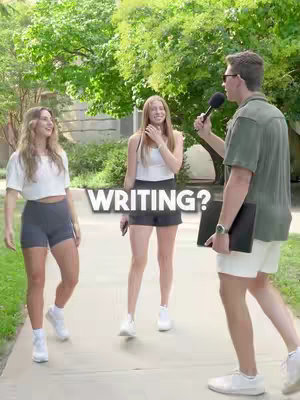 A group of three young people, two women and one man, are standing outside on a walkway. The man holds a microphone towards the two women as if he is conducting an interview. The woman on the left faces the camera and the woman in the middle is looking at the man. A text overlay, ?WRITING?, is positioned in the center of the frame near the individuals. The background includes green trees and a stone walkway.