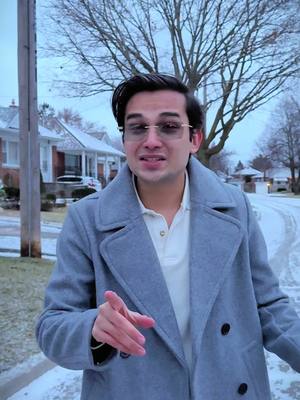 A man wearing a grey coat and glasses stands outdoors on a snow-covered road, pointing his finger. Houses and leafless trees are visible in the background against a cloudy sky. The scene appears to be filmed in a residential area.