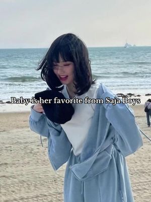 A young woman stands on a beach with a slight smile, holding a black hat. She wears a light blue jacket and pants over a white top. The background shows the ocean and distant land.