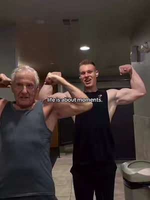 Two men flexing their biceps in a gym-like setting. The older man is on on the left, wearing a gray tank top, while the younger man is on the right, wearing a black tank top with the Gymshark logo. Text "life is about moments." is overlaid at the bottom. The background shows what seems to be a gym. 