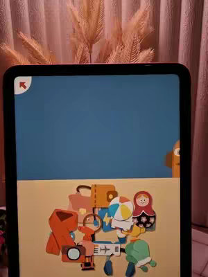 An iPad screen displays a colorful puzzle game. Various cartoon-style objects are arranged on a sand-colored surface, with a blue background above. In the very top, there's a reddish-brown curtain and some dried flower stems.