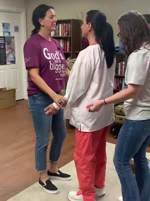 Three women are standing in a room, seemingly engaged in a spiritual practice. The main figure in the center is wearing a white lab coat and red pants. Two other women are facing her, one is wearing a purple shirt and blue jeans, and the other is wearing a plain white shirt and jeans. They are in a well-lit room with bookshelves in the background. The overall composition suggests a scene focused on faith or religious practice.