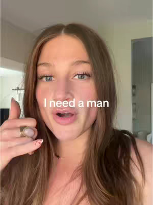 A selfie-style shot of a woman with long, light brown hair, angled slightly to the right. The background is a soft, blurred indoor setting with hints of green and white. Text overlay 'I need a man' is positioned above the woman's mouth. She has blue eyes, and her mouth is slightly open as if she is speaking. A ring is on her left ring finger. The overall composition is well-lit and focused on the woman's face.