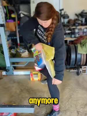 A woman in a black sweatshirt and leggings stands in what appears to be a garage or storage space. She holds a yellow folder and book in her hands, looking down at them. The hook text 'anymore' is added as a yellow label at the bottom of the image.