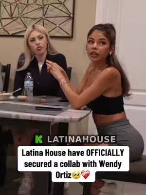 Two young women are seated at a table indoors. One is blonde, wearing a black top, and looking inquisitively to her left. The other has dark hair and is wearing a black sports bra and grey shorts, gesturing with her left hand. Text overlaid on the image is a lower third.