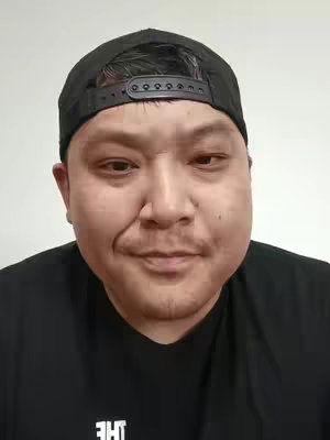A close-up shot of a man wearing a black baseball cap, facing the camera directly. He has a neutral expression. He is wearing a black shirt, and the background is a plain, light color.