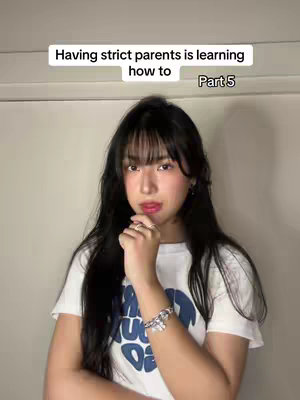 A young woman with long black hair and bangs is positioned in front of a plain white wall. She is wearing a white t-shirt with a graphic design and has some bracelets on her wrist. The text overlay at the top of the image reads, "Having strict parents is learning how to Part 5". She's holding her index finger near her mouth, as if contemplating or in thought.