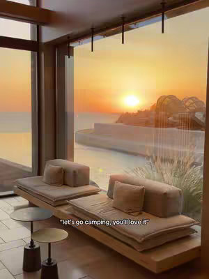 The image captures a luxurious indoor setting overlooking a sunset. Through a large window, the viewer sees a vast ocean and a setting sun painting the sky in warm oranges and yellows. Inside, a modern seating arrangement is visible, featuring a wooden platform with cushions and pillows. Small side tables and an overall minimalist design contribute to the serene, high-end vibe of the space. The image composition makes use of natural light and perspective, which emphasizes the beautiful outdoor scenery displayed through the oversized window.