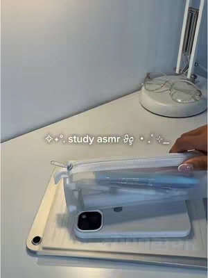 The cover image shows a close-up shot of items on a white desk. A transparent pencil case containing pens is resting on top of a white iPad-like device. The case is slightly angled towards the camera. The background includes a blank white wall and a portion of a desk lamp and a set of glasses. The image utilizes cool tones with a slight aesthetic filter.