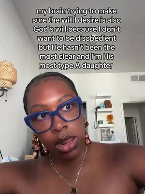 A selfie of a woman with dark skin and short, dark hair and braids. She's wearing blue-rimmed glasses and has a serious expression. Text overlay is above her head and partially obscuring her face. The background shows a well-lit room, with a lamp and a shelf with decorations.