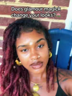 A selfie-style shot featuring a dark-skinned woman with red-tinted locs, gold jewelry, and facial piercings. The background includes a brick wall and a blue chair, suggesting an outdoor setting. Text overlays the top of the image.