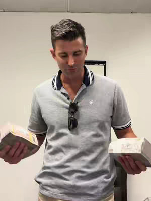 A man in a gray polo shirt stands in a room, holding stacks of cash in both hands. The background is a plain white wall, and a dark-framed mirror is partially visible behind him. His sunglasses are hanging on his shirt. He appears to be looking down.