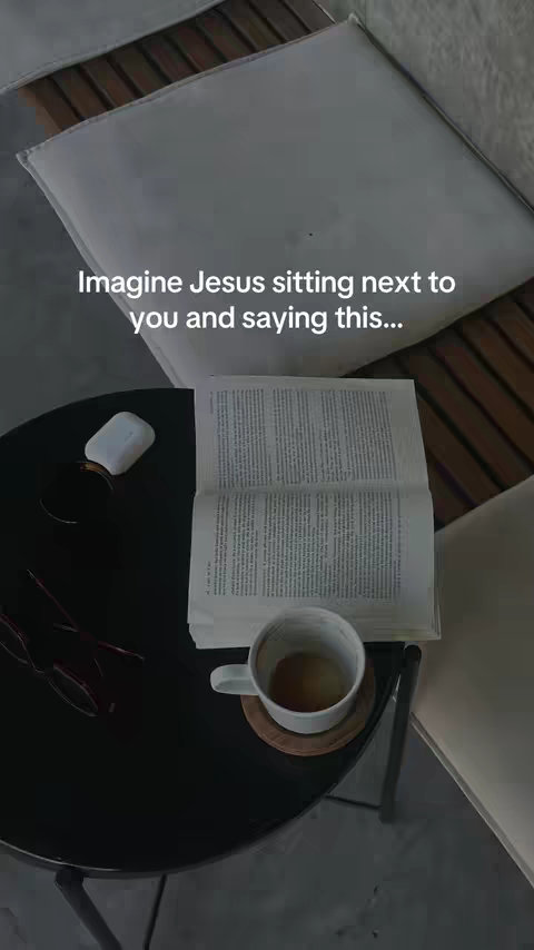 An overhead shot of a black table with objects on it in focus: a coffee cup, an open book, a small container, and what appears to be a pair of glasses. The background includes a section of a wooden chair and a portion of the seating.