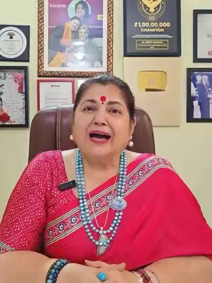 A woman with fair skin, wearing a red sari with a jeweled necklace, holding a microphone with a background of frames and a sign.