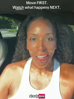 A woman with dark skin is in a car, looking forward, with a white tank top and gold jewelry. The sunlight is bright and the background is blurred.