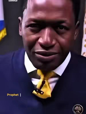 A man in a dark suit and yellow tie is the central focus. The man has dark skin and a serious expression, looking towards the viewer. The background is indistinct, with only glimpses of flags in the upper left. The text overlayed at the bottom reads 'Prophet'.