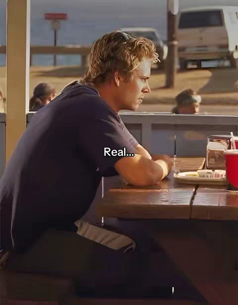 A man with light-brown hair sits at a table on a sunny day, looking pensive. The background includes a beach scene and a white van.