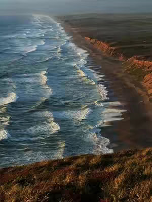 An aerial view of a coastline with a beach and ocean. The sun is setting, casting a warm glow on the scene. The image is dominated by the ocean on the left, with waves breaking near the shore. The beach runs along the right side, and the coastline curves gently into the distance. The colors are muted, with blues, browns, and oranges.