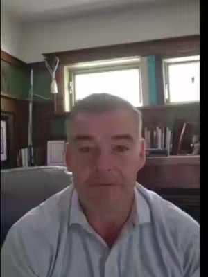 A middle-aged man with short, gray hair sits in a room and looks at the camera. He's wearing a light blue button-down shirt. The background includes a dark wooden wall, a window, and a hint of a bookshelf.