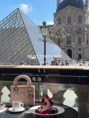 A cityscape in Paris featuring the Louvre Pyramid and the Louvre Museum in the background. In the foreground, a table is set with a pink handbag, a cup of espresso, and a dessert with whipped cream and berries. A glass railing and a cafe setting are present.