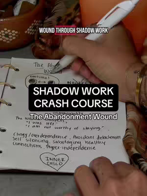The image shows someone's hand holding a pen and writing on an open notebook. The notebook's pages display handwritten notes and diagrams related to shadow work and The Abandonment Wound, with a black box overlay containing text that reads 'SHADOW WORK CRASH COURSE'.