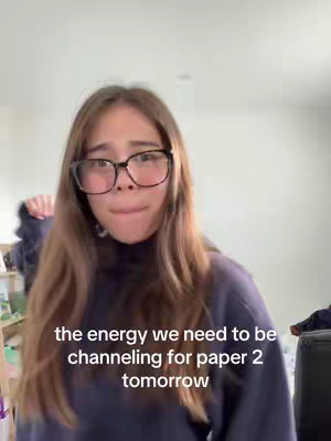 The cover image features a young woman looking directly at the camera with a somewhat worried or stressed expression, her mouth slightly open. She is wearing glasses and a dark-colored top likely a turtleneck, with long brown hair. The background is a simple, light-colored wall. The text overlay says "the energy we need to be channeling for paper 2 tomorrow".