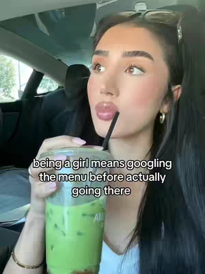 A woman is seated inside a car, holding a clear plastic cup filled with a green beverage and a black straw. Her lips are glossy, and she's gazing off to the side. Text overlays the image, and the background shows more of the car's interior.