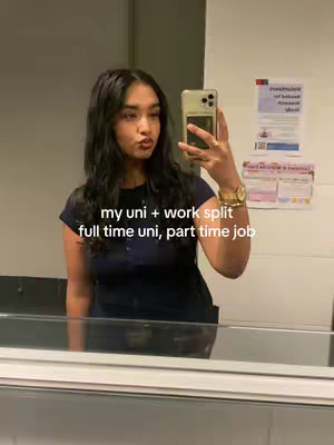 A woman is taking a selfie in a bathroom mirror, holding a gold-colored phone. She has long, dark hair and is wearing a dark blue top. Text overlays the image: 'my uni + work split' and 'full time uni, part time job'.